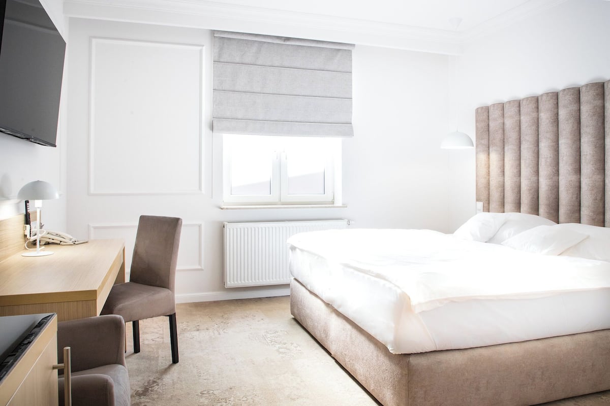 A hotel room with a bed, a desk with a chair, a television, and a window with a blind.