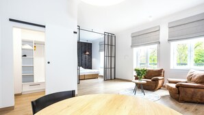 A modern living room with a wooden dining table, a sofa, and a chair. There is a large window with curtains, a bed with a headboard, and a wardrobe with a mirror. - Europejski Boutique Hotel (Radom)