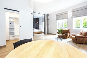 Una sala moderna con una mesa de comedor de madera, un sofá y una silla. Hay un ventanal con cortinas, una cama con cabecero y un armario con espejo.