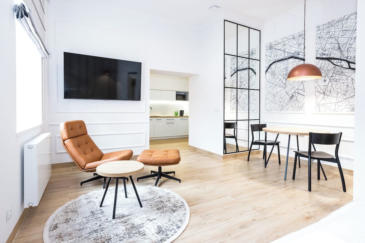 A modern living room with a flat-screen TV, a dining area with a round table and chairs, and a large window with a cityscape view.