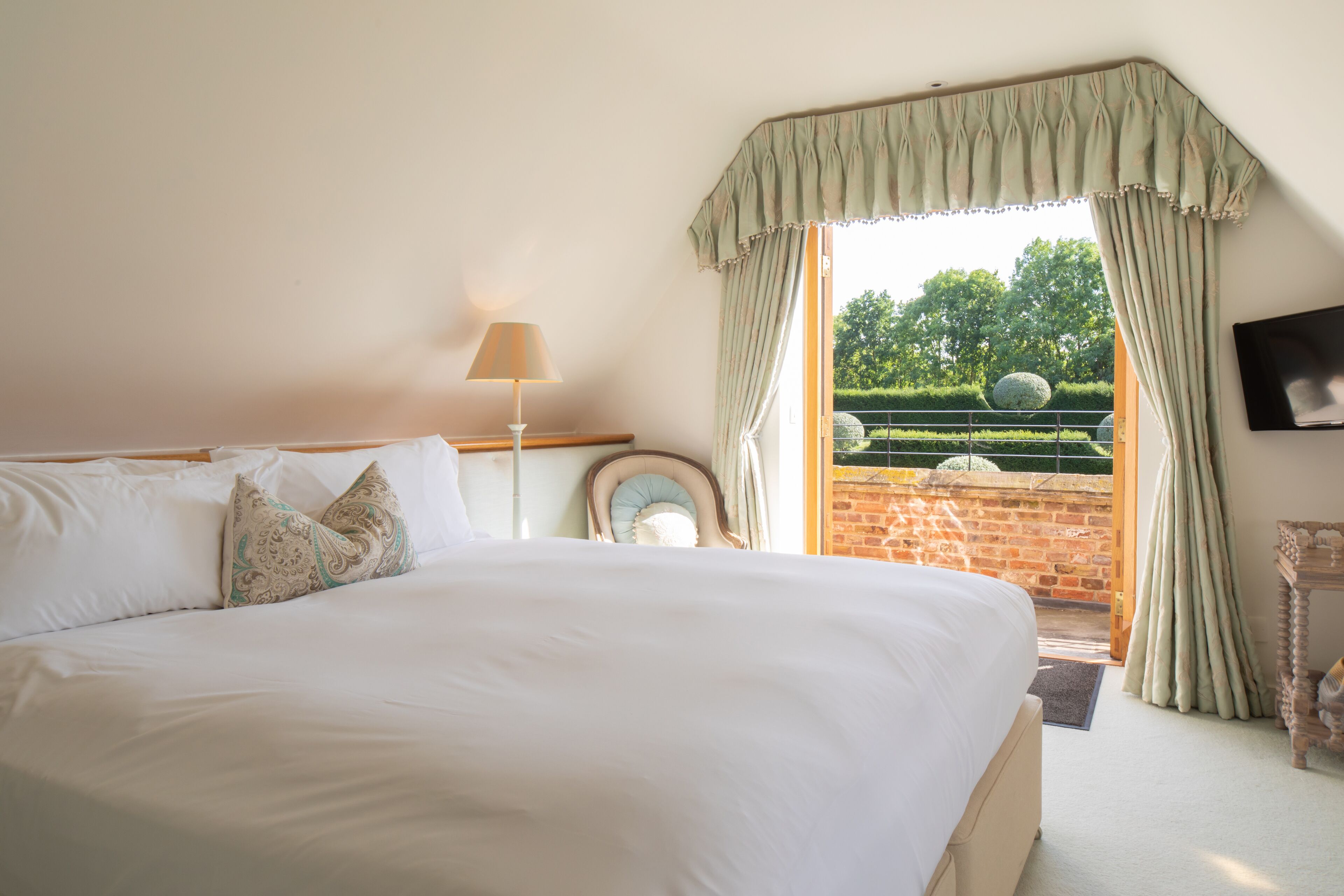 A bedroom with a large bed, a bedside table with a lamp, a mirror, and a view of the garden through open curtains.