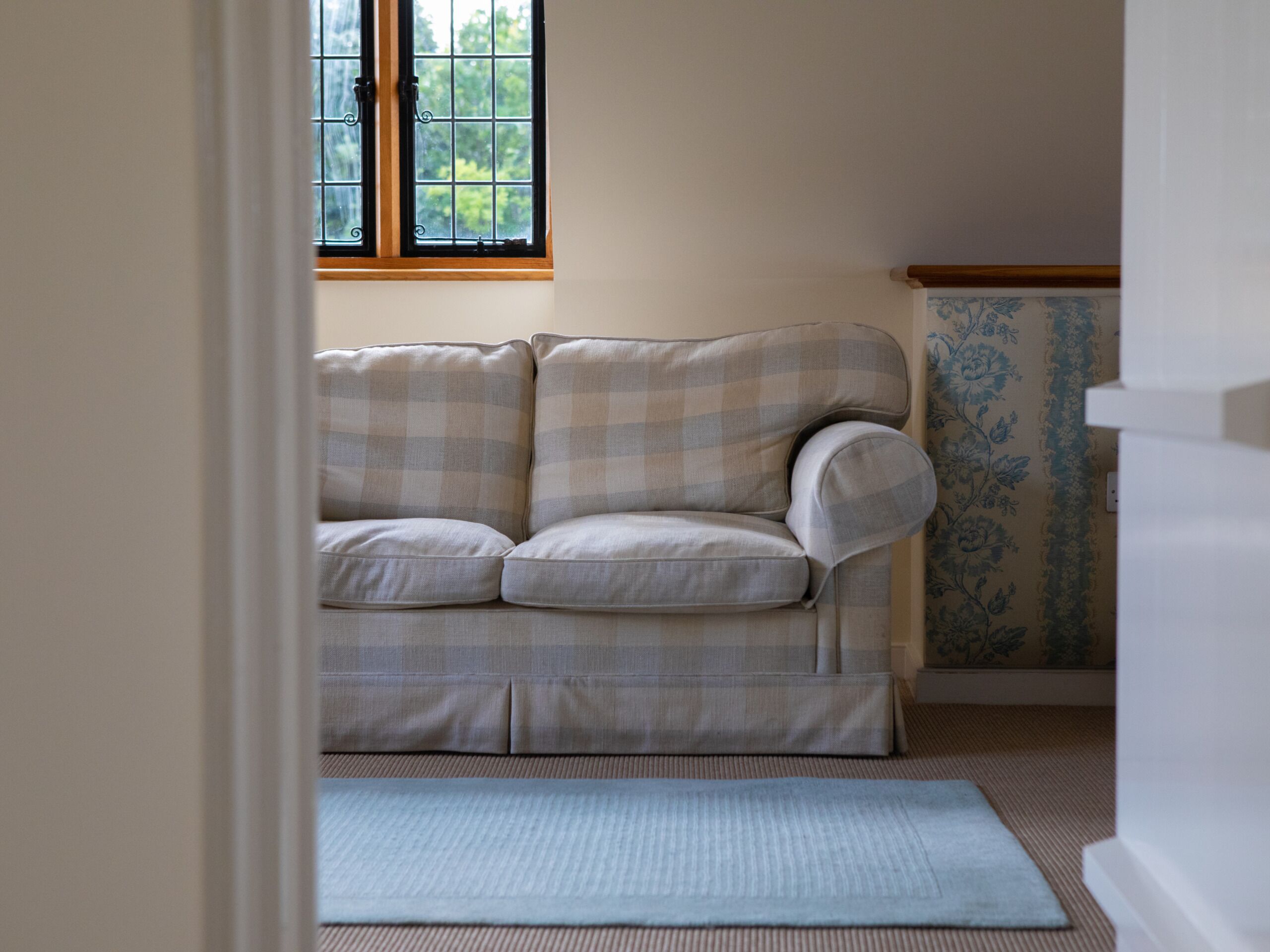 A room with a beige checkered sofa, a window with a view of greenery, and a patterned wallpaper on the wall.
