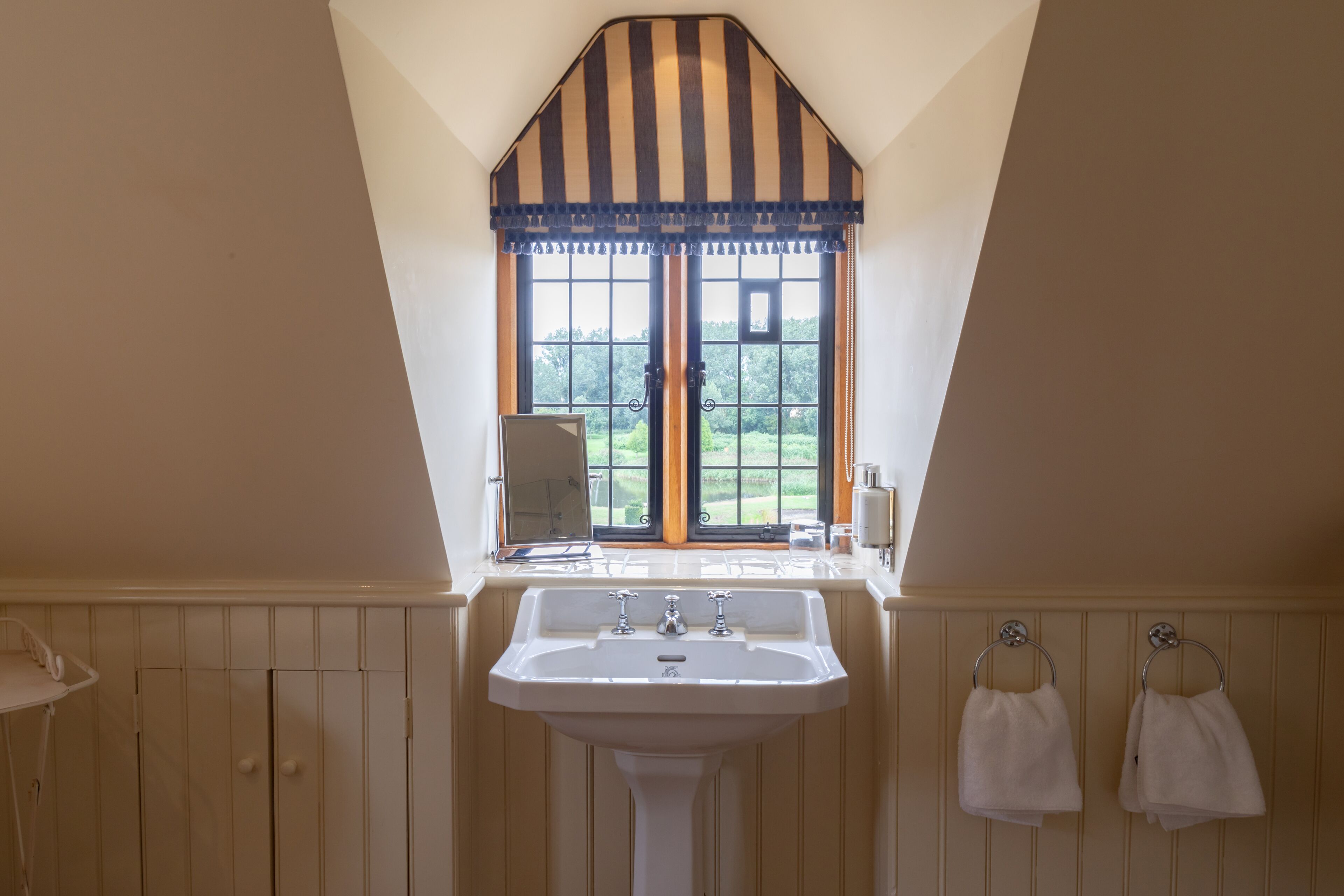 A bathroom with a white sink, wooden paneling, a window with a striped valance, and two towels hanging on hooks.