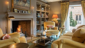 Un salon chaleureux avec une cheminée, des fauteuils confortables, une bibliothèque et une vue sur l’extérieur.