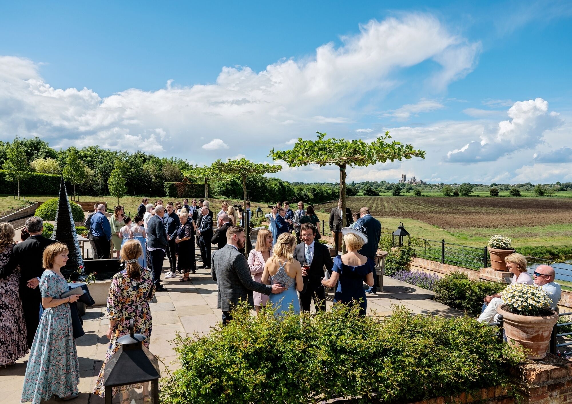 A group of people gathered outdoors, possibly at a social event, with a clear sky and greenery in the background.