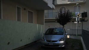 A car parked in a narrow driveway with a tree and a building in the background. - Residenza Manuela (Riccione)