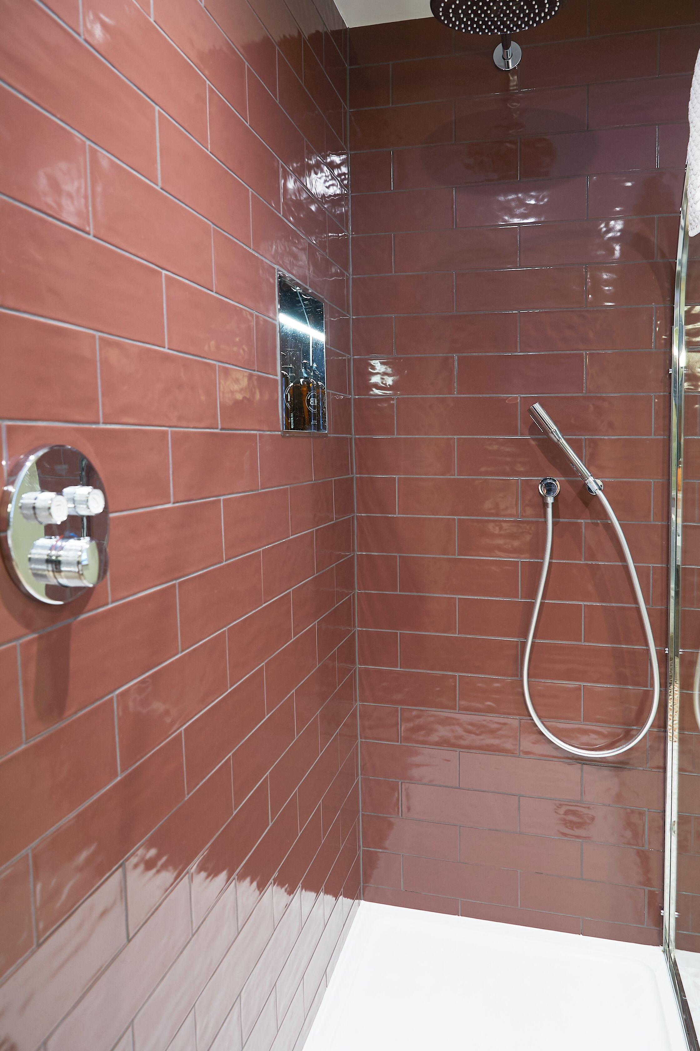Ein Badezimmer mit roten Fliesen an den Wänden, einer Dusche mit Duschhandbrause und einem wandmontierten Wasserhahn.