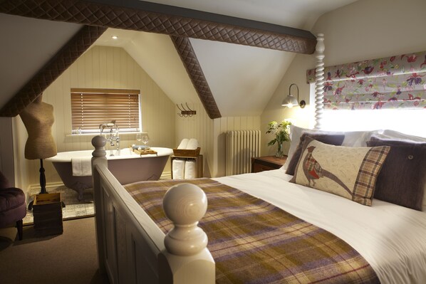 A bedroom with a large bed, a window with floral curtains, a built-in bathtub, and a wooden ceiling. - The Plough Scalby (Scarborough)