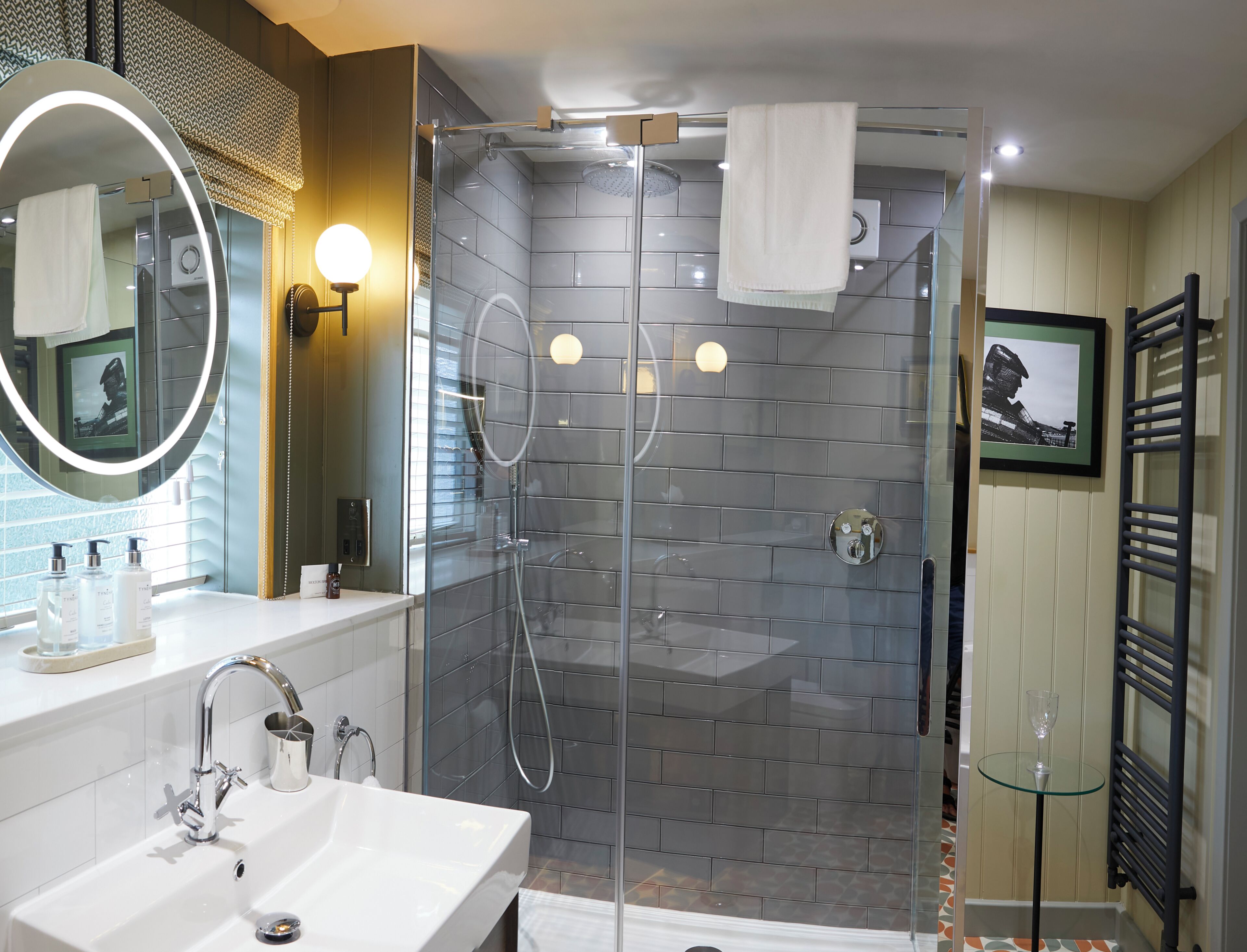 Un baño moderno con ducha envuelta en vidrio, lavamanos blanco y un espejo grande con luz circular.