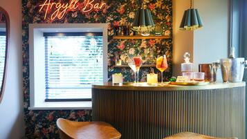 A bar area with a floral wallpaper, a neon sign reading 'Argyll Bar', and a window with blinds. There are two bar stools and a variety of drinks on the counter.