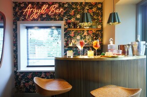 A bar area with a floral wallpaper, a neon sign reading 'Argyll Bar', and a window with blinds. There are two bar stools and a variety of drinks on the counter.