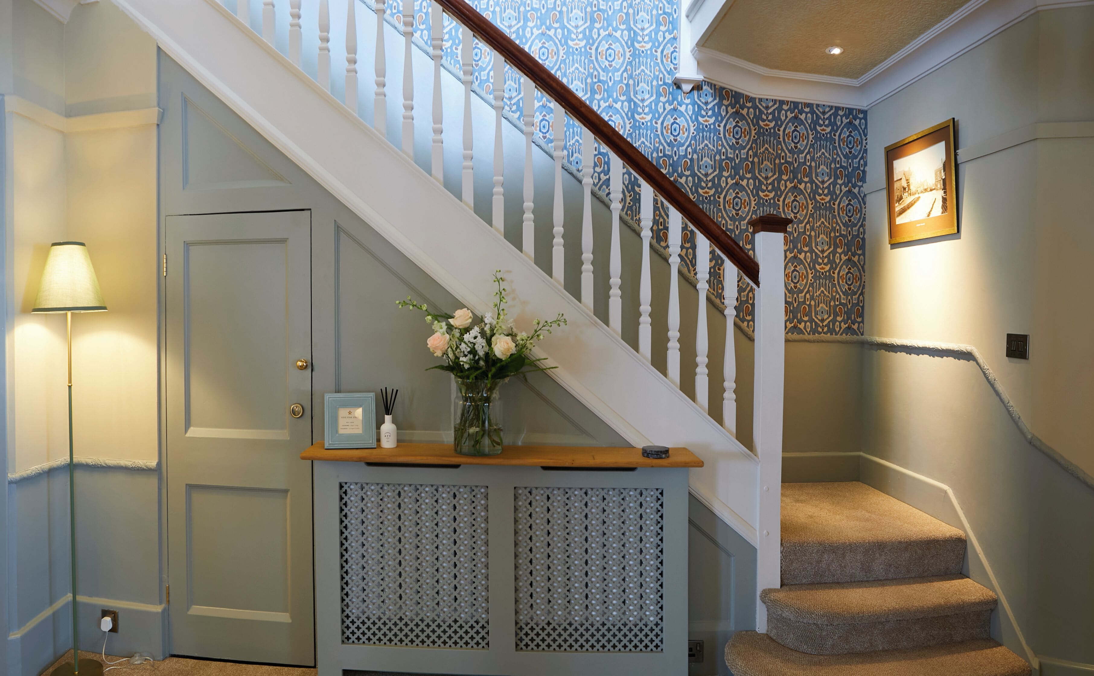 A staircase with a patterned wall, a door with a light switch, a lamp, and a small table with a vase of flowers.