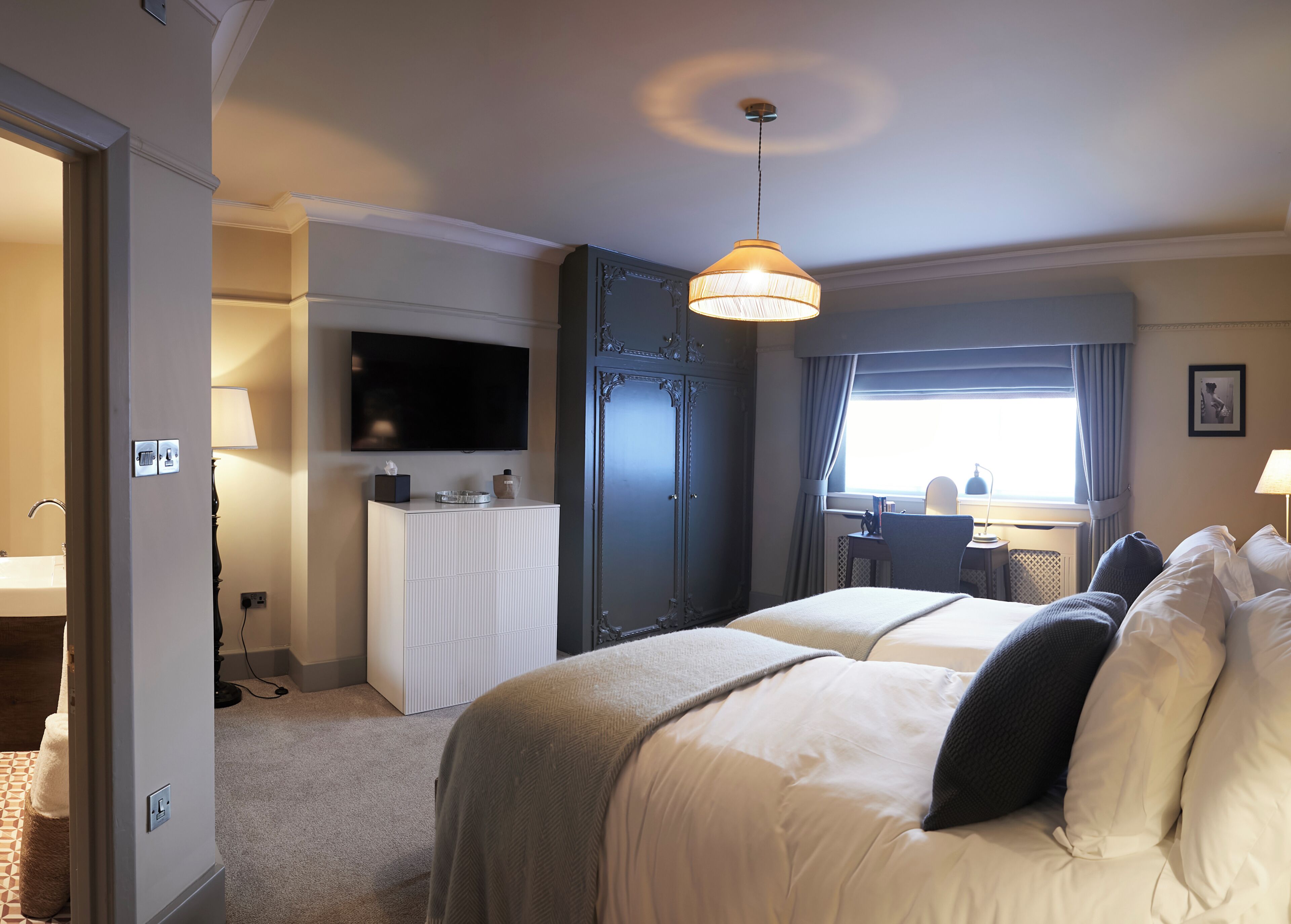 A hotel room with a bed, bedside table, lamp, TV, wardrobe, and a window with curtains.