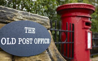Un cartel con la inscripción 'THE OLD POST OFFICE' sujeto a una pared de piedra, con un buzón rojo y una valla metálica al fondo.