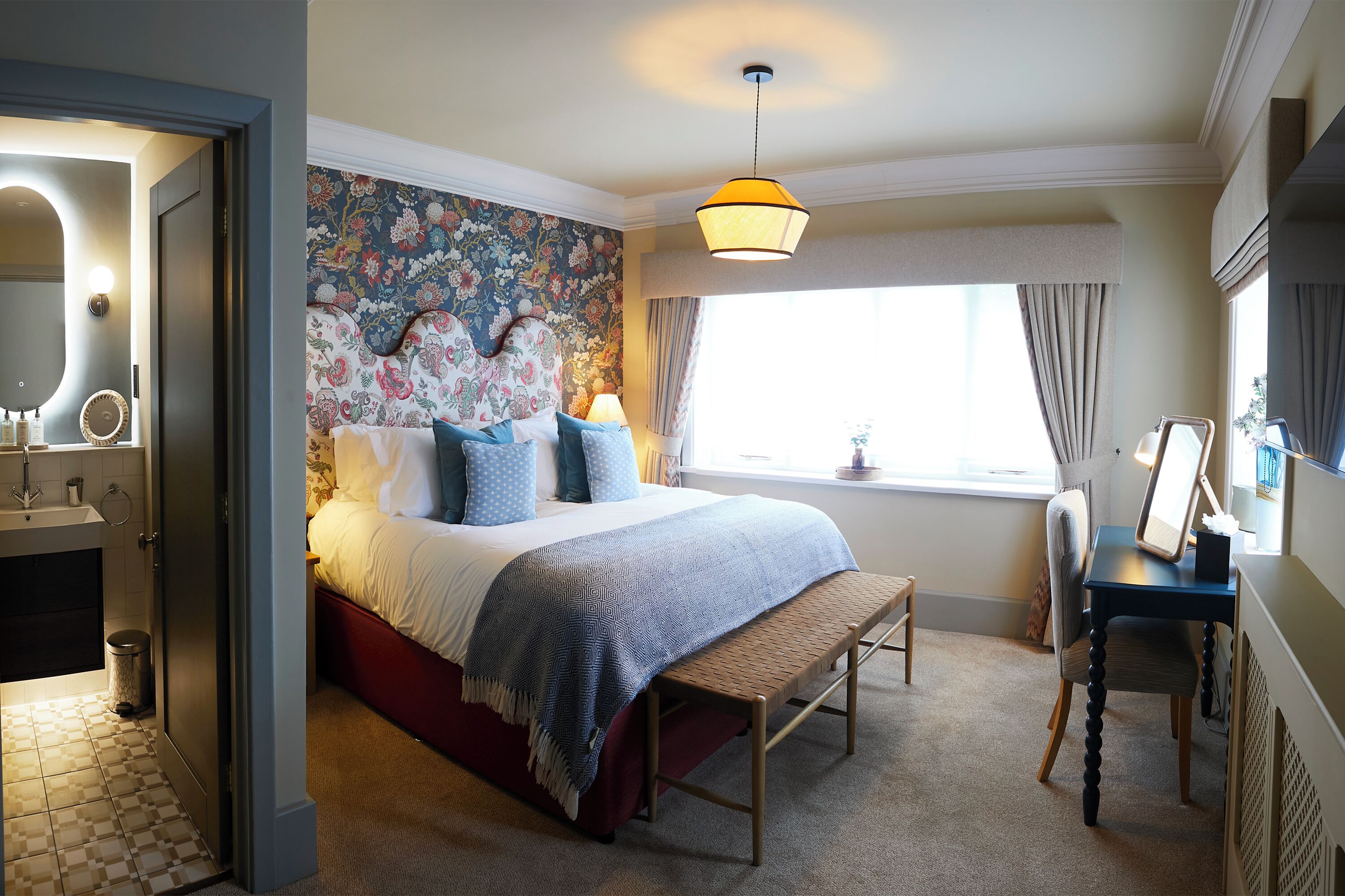 A hotel room with a large bed, a bedside table, a window with curtains, a bathroom with a mirror, and a wall with a floral pattern.