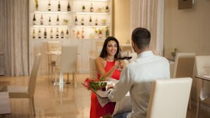 Um casal jantando à mesa com uma garrafa e um copo de vinho, em um restaurante com adega ao fundo.