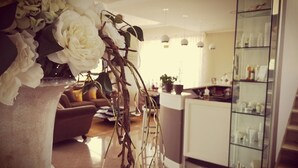 Uma sala de estar moderna com um vaso grande de flores brancas, um sofá e um aparador de vidro com diversos objetos.