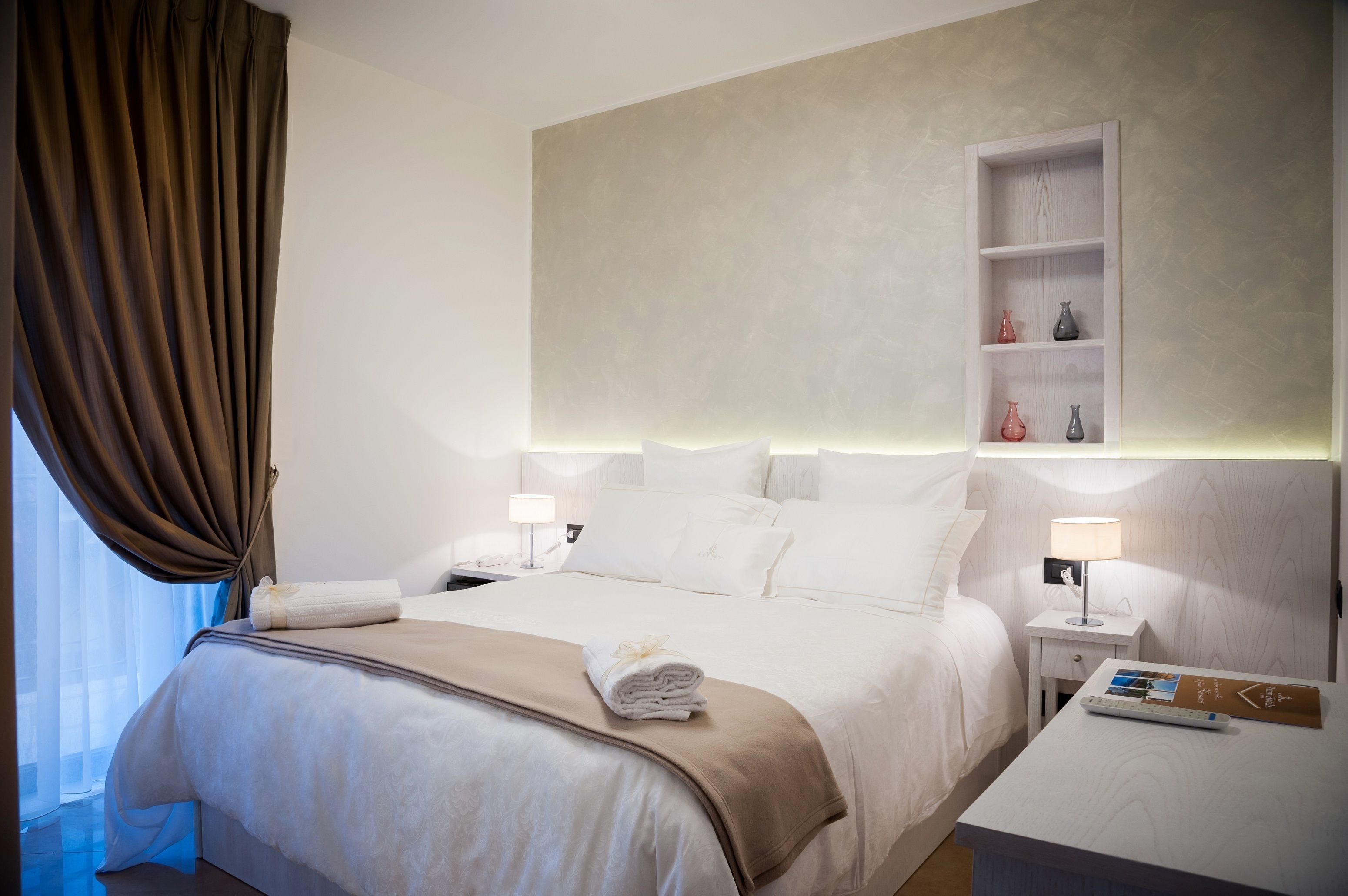 Una camera d'albergo con un letto grande, due comodini, un piano d'appoggio, uno scaffale con oggetti decorativi e una finestra con tende.
