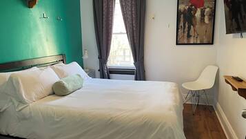 Una cama bien hecha con sábanas blancas y una única almohada verde, una silla blanca y un cuadro de una pareja en la calle.
