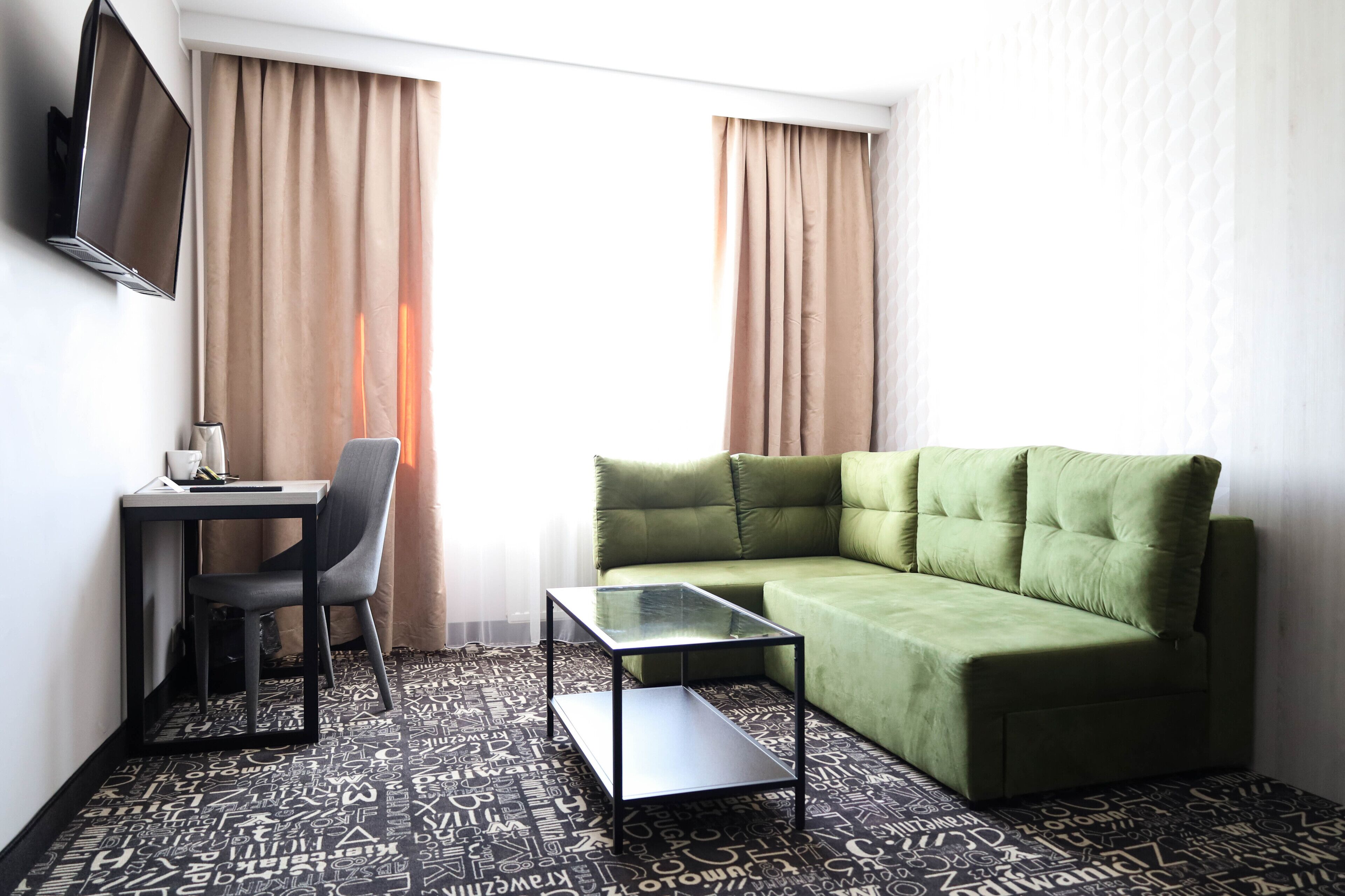 A room with a green sofa, a glass-top coffee table, a black chair, and a TV mounted on the wall.