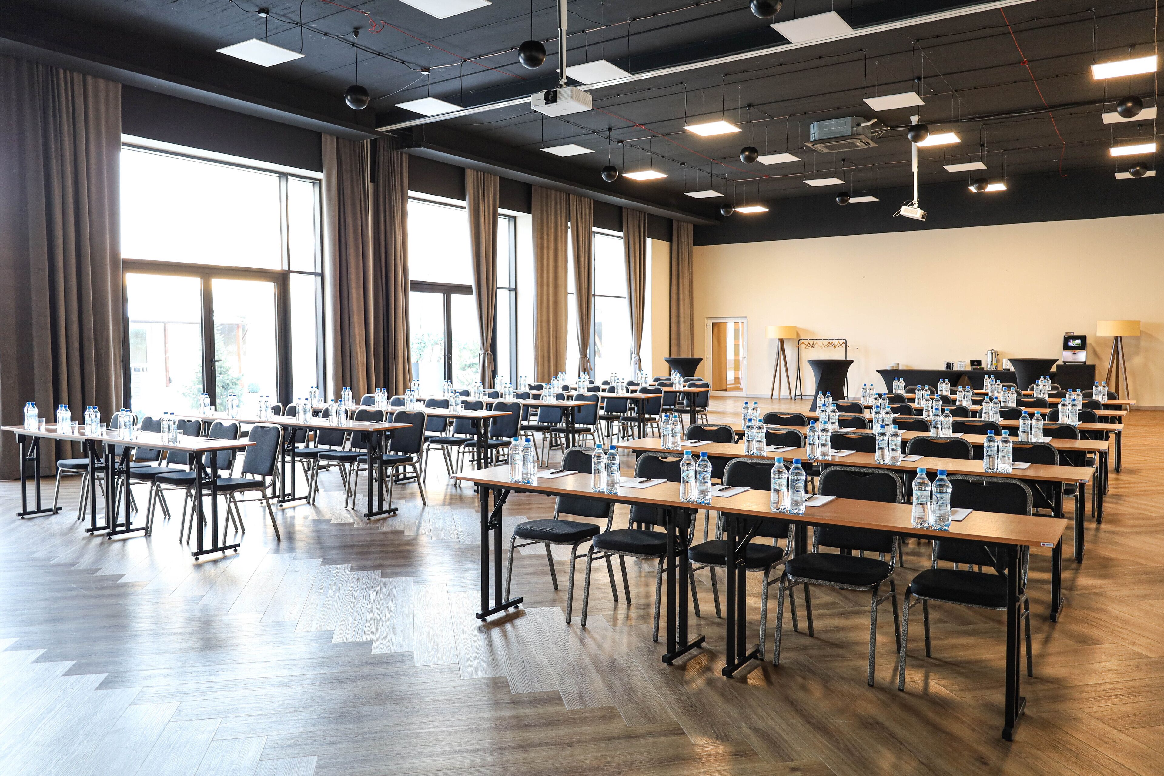 Un salón de conferencias con varias mesas y sillas, equipado con botellas de agua y proyectores.