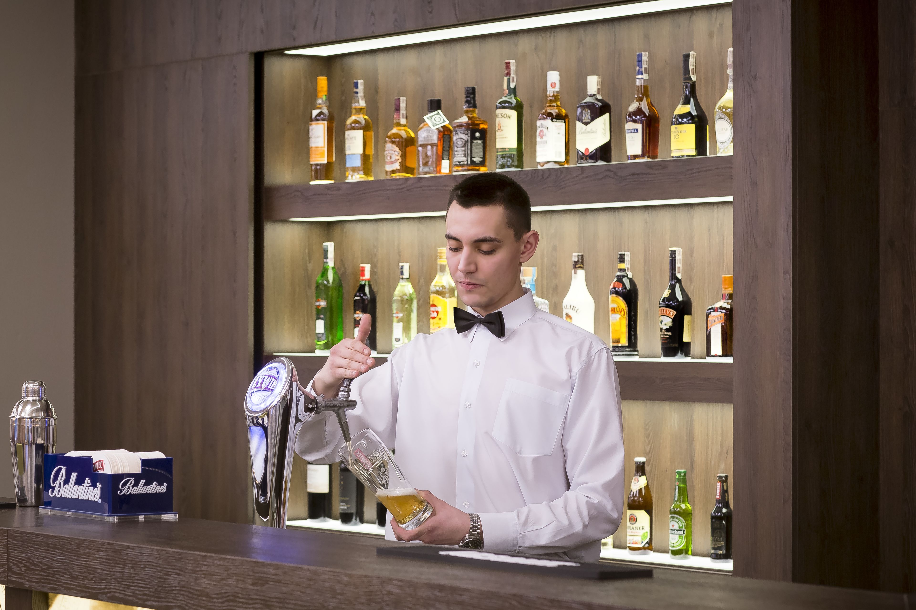 Uma área de bar com um bartender servindo uma bebida, prateleiras com diversas garrafas de licor e uma caixa de cerveja Budweiser no balcão.
