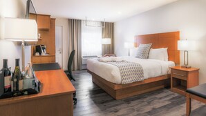 Quarto de hotel com cama de madeira, escrivaninha com cadeira, mesa de cabeceira e abajur. Há garrafas sobre a escrivaninha.