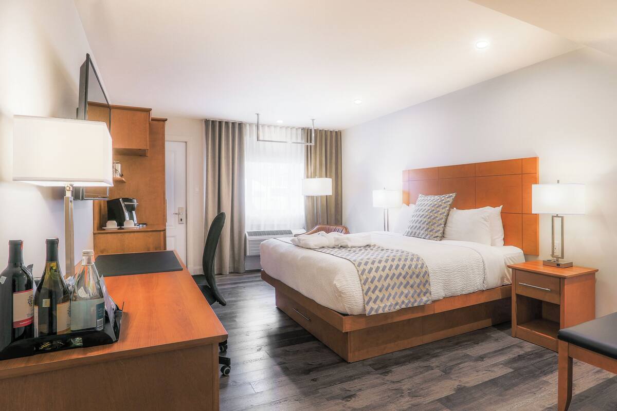 A hotel room with a wooden bed, a desk with a chair, a nightstand, and a lamp. There are bottles on the desk.