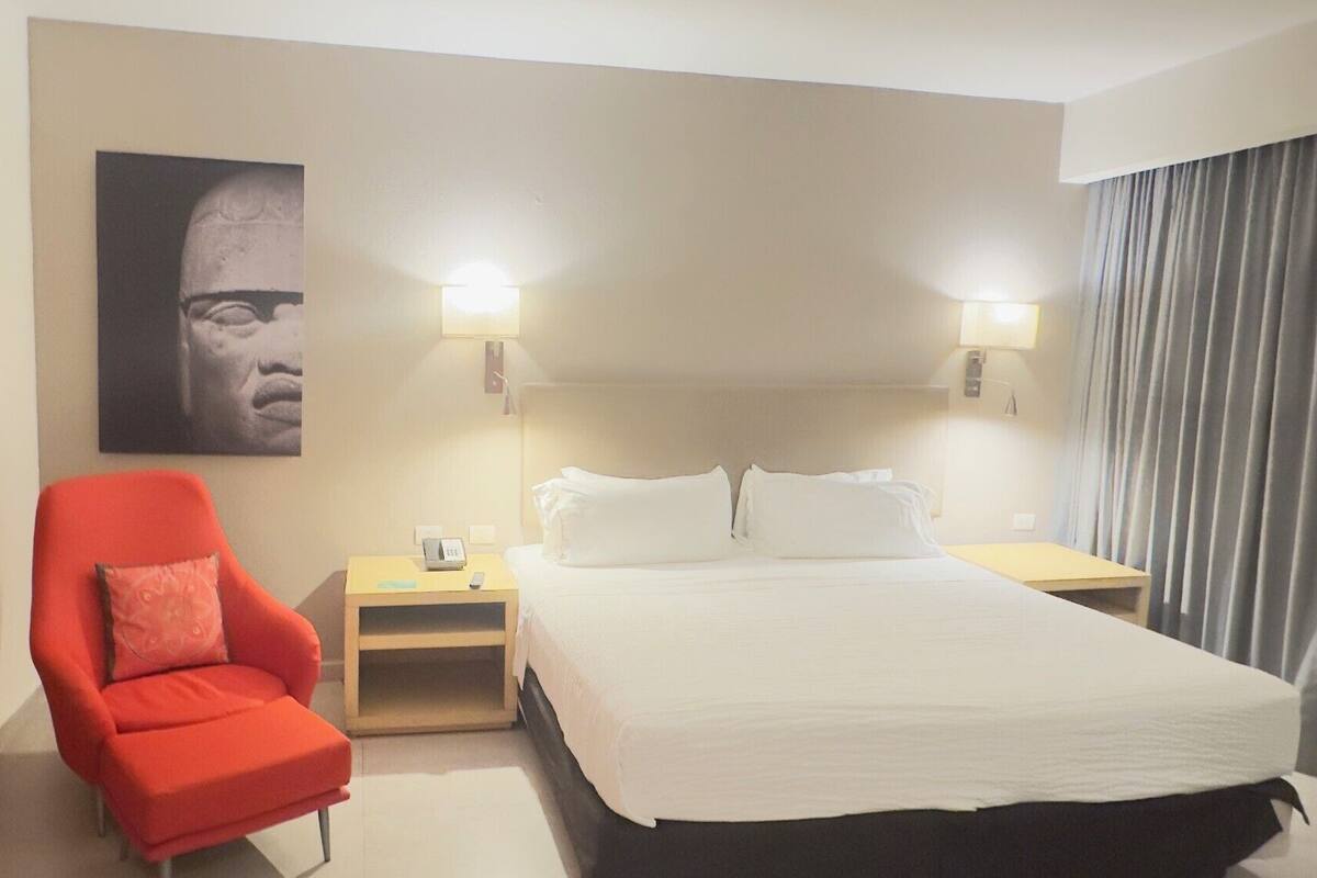 A hotel room with a bed, a red armchair, a nightstand with a lamp, and a wall-mounted picture.