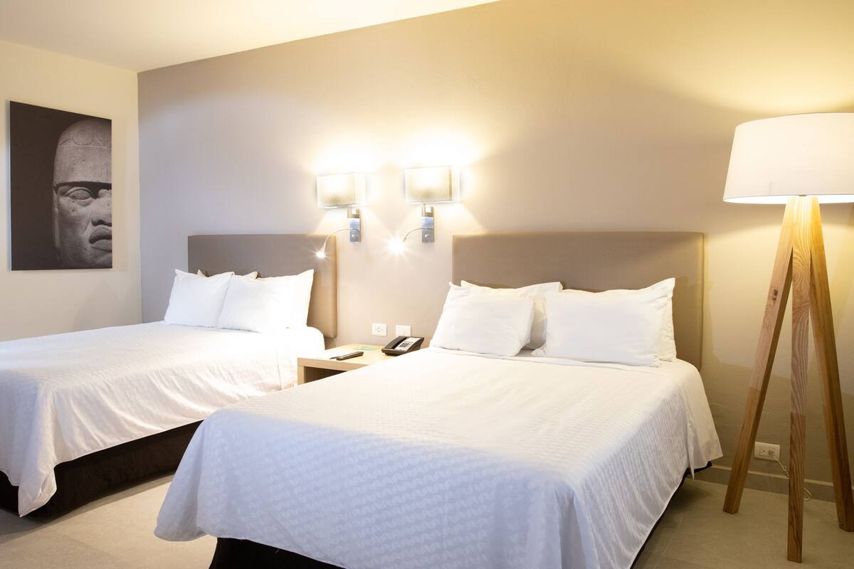 A hotel room with two beds, a nightstand with a phone, wall-mounted lights, and a standing lamp.