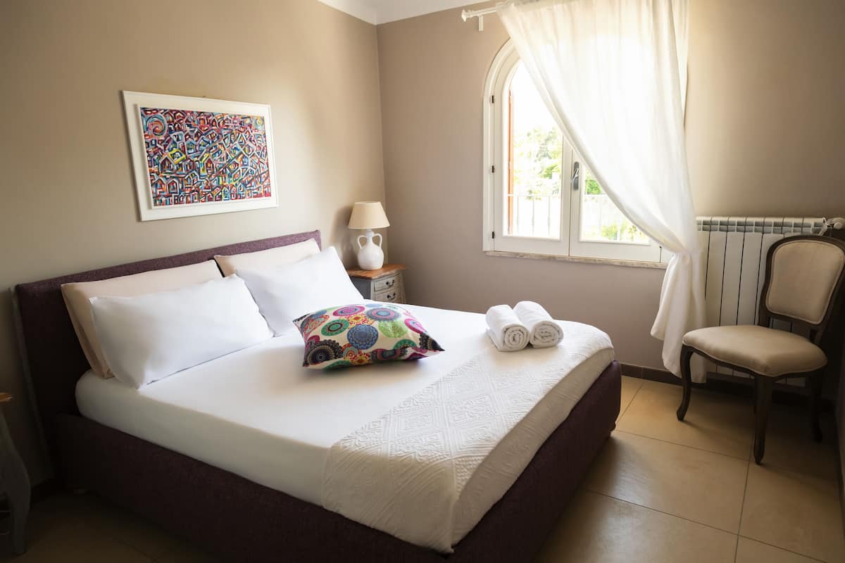A neatly made bed with white linens, a colorful pillow, and rolled towels on a headboard. There is a chair to the right and a window with curtains.