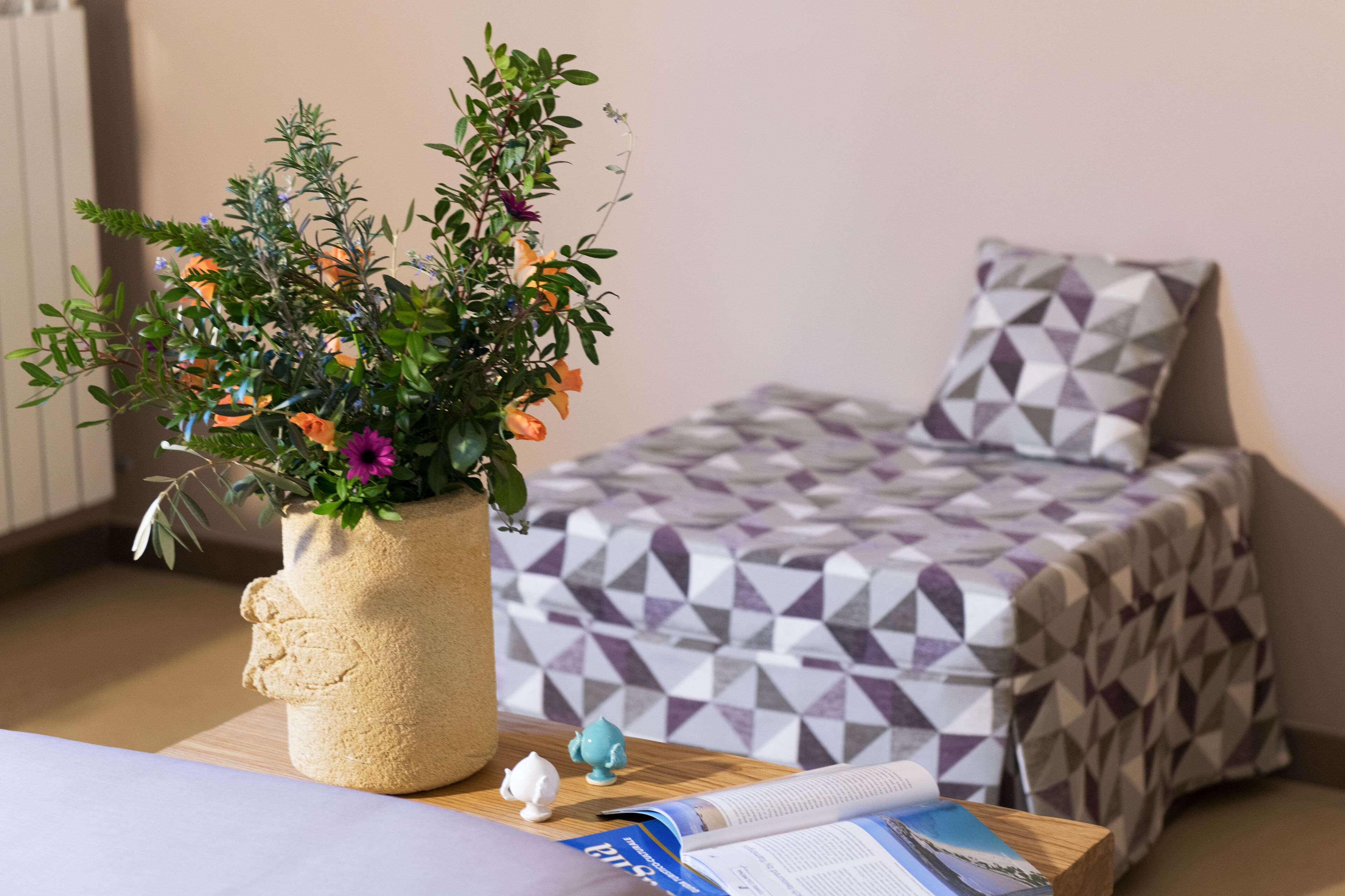 Un letto rifatto con cura, con una coperta a motivi geometrici, una piccola pianta in vaso con fiori vari e un comodino di legno con un libro e una piccola statuina.
