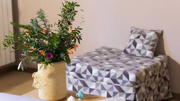 Un letto rifatto con cura, con una coperta a motivi geometrici, una piccola pianta in vaso con fiori vari e un comodino di legno con un libro e una piccola statuina.