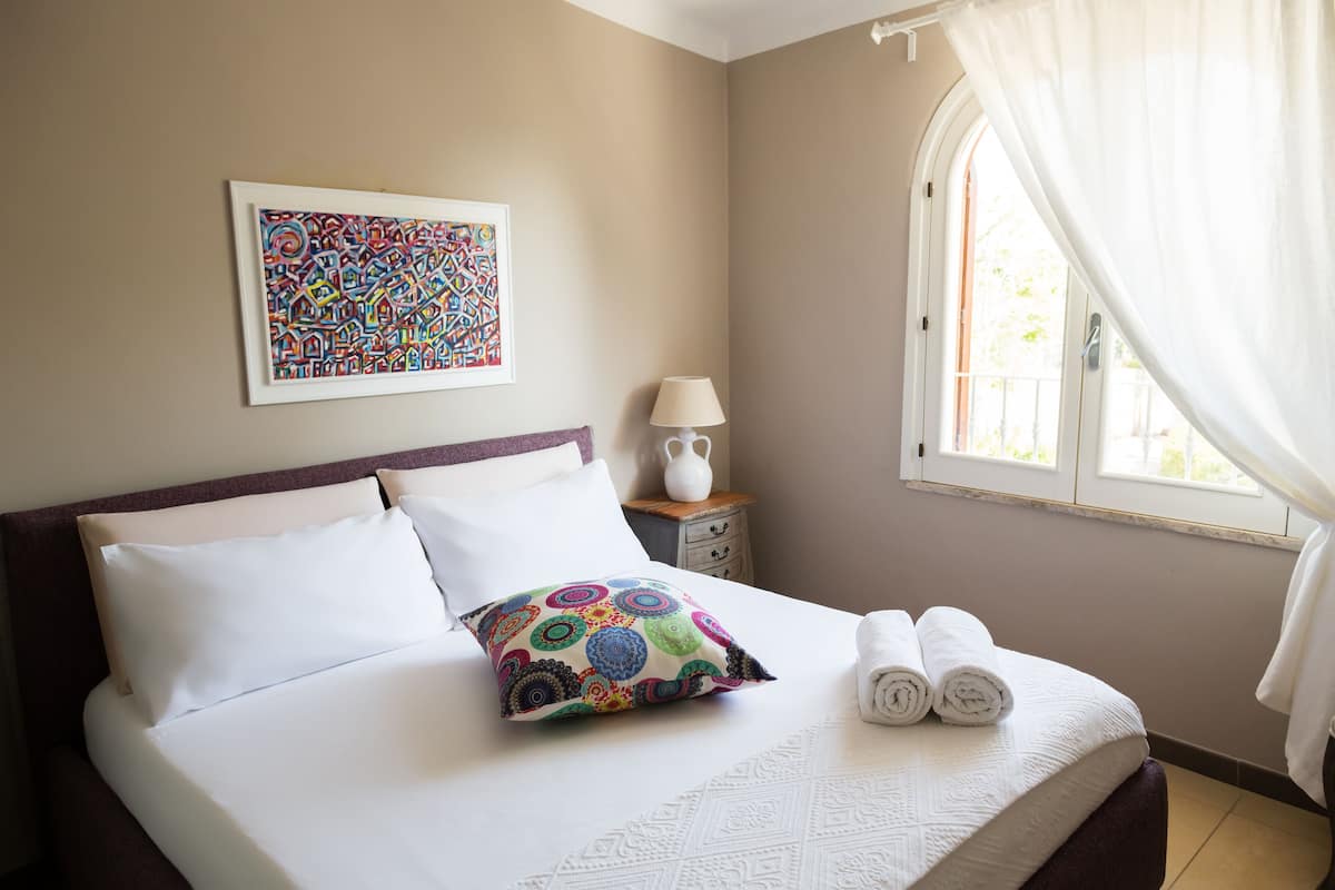 A neatly made bed with white linens, a colorful pillow, and rolled towels.