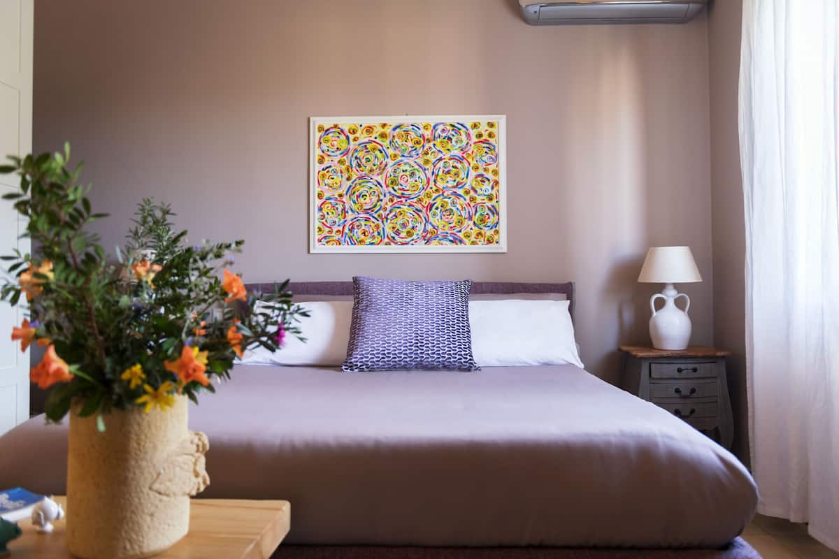 A bedroom with a bed, a bedside table with a lamp, a decorative vase with flowers, and a colorful abstract painting on the wall.