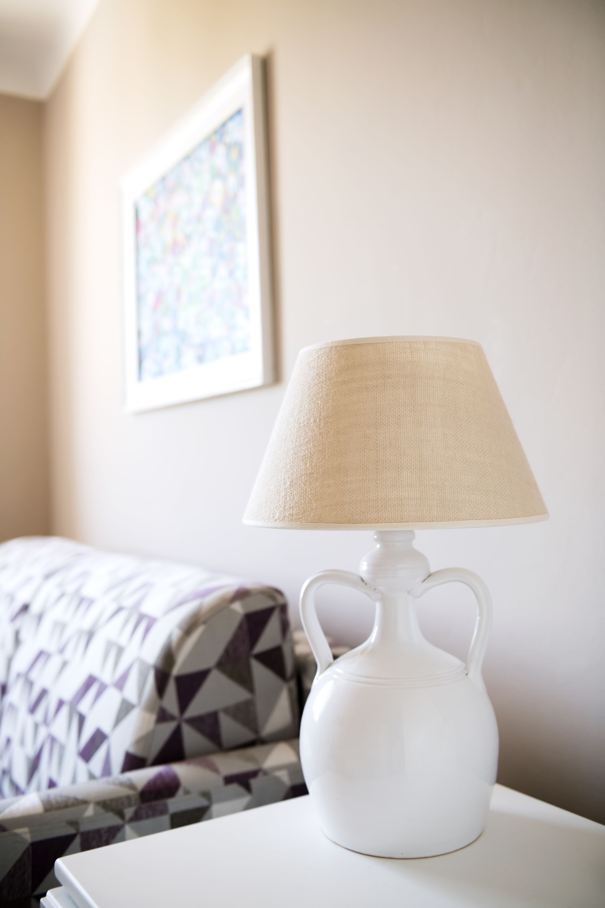 A white table lamp with a beige lampshade on a white table in a hotel room.