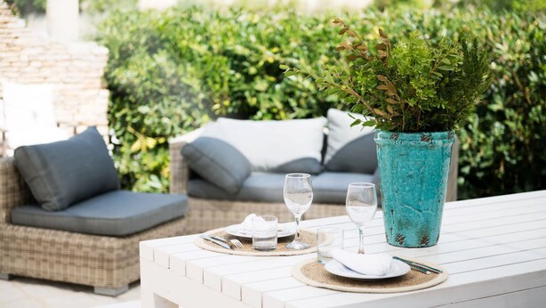 Un área de comedor al aire libre con una mesa blanca, dos cojines grises, un florero turquesa con follaje y un sofá de mimbre al fondo.
