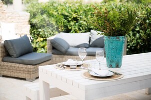 Un área de comedor al aire libre con una mesa blanca, dos cojines grises, un florero turquesa con follaje y un sofá de mimbre al fondo.