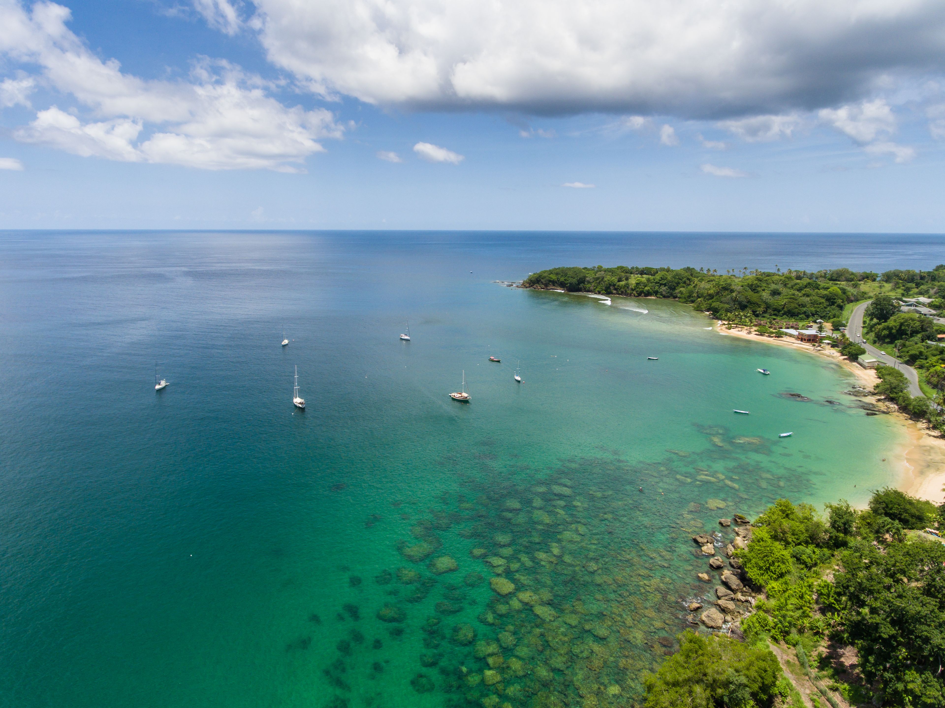 An aerial view of a coastal area with clear turquoise waters, several sailboats, and a beach with lush greenery.