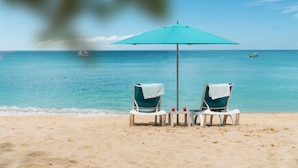 Two beach chairs under a blue umbrella on a sandy beach with the ocean in the background.