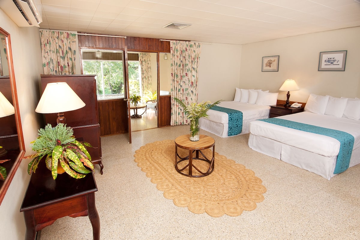 A hotel room with a bed, bedside table, lamp, potted plant, and a view of the outdoors through a window.