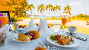 A table set for breakfast with plates of food, glasses of water and juice, and a view of palm trees and the ocean.