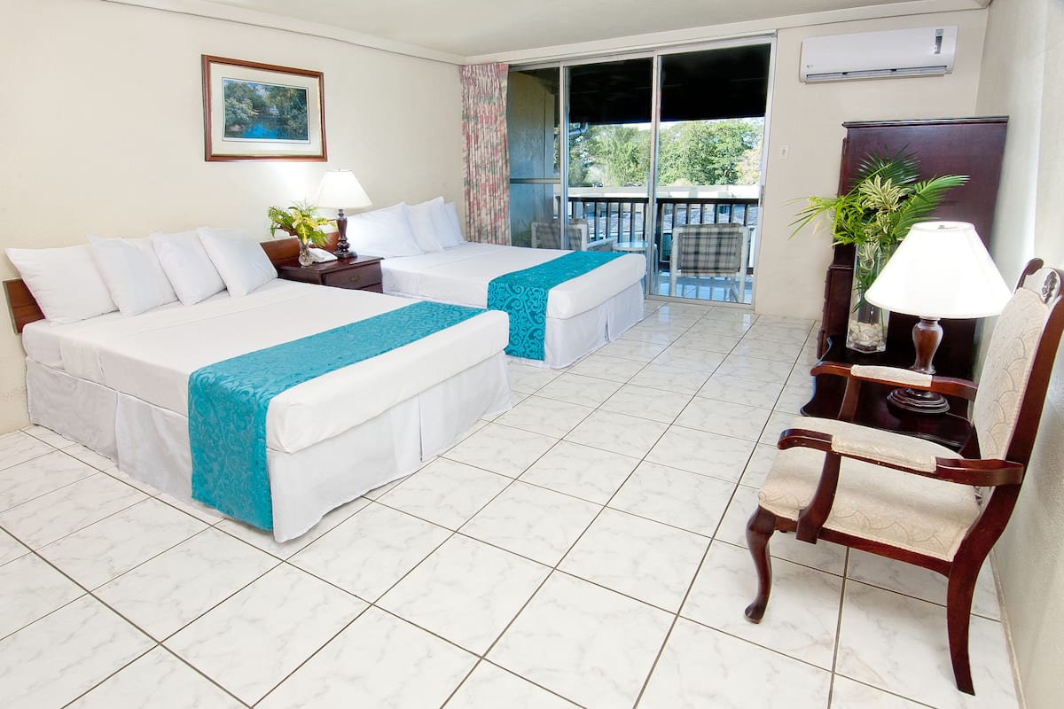 A hotel room with two beds, a chair, a table, a plant, and a balcony with a view.