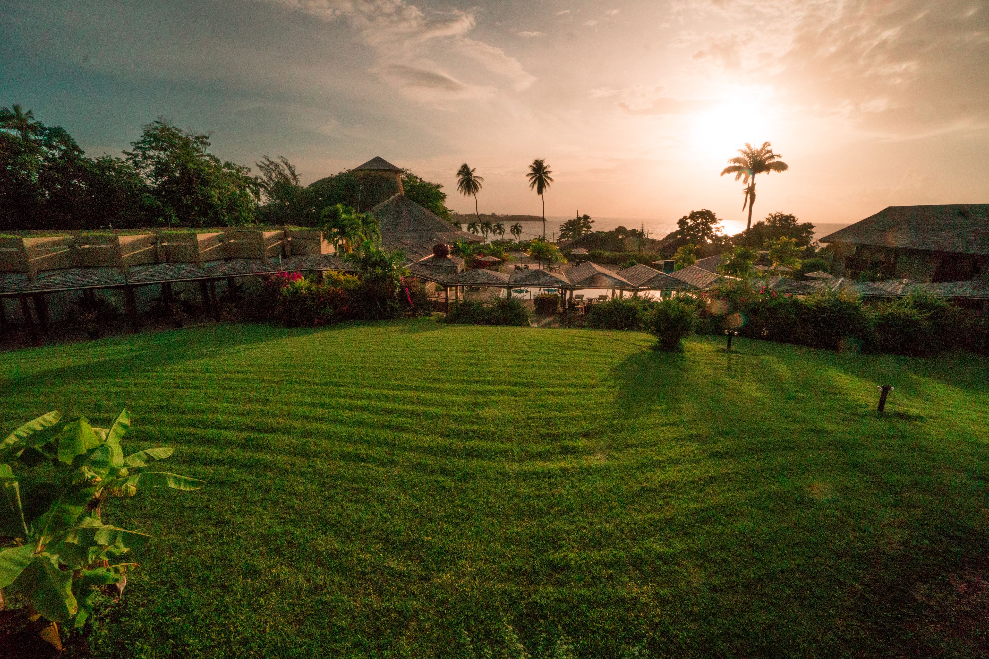 A resort with a well-maintained lawn, several thatched-roof structures, and a view of the ocean at sunset.