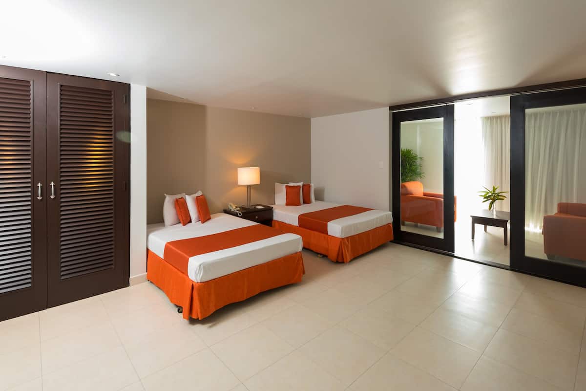 A hotel room with two beds, a desk, a chair, a lamp, and a sliding glass door to a balcony.