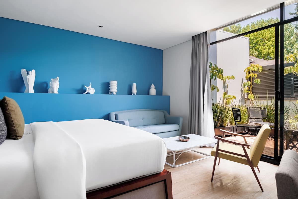 A modern bedroom with a blue accent wall, a bed, a sofa, a coffee table, and a view of the outdoors.