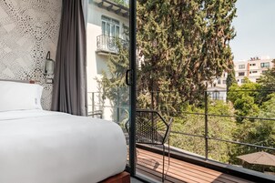 Un dormitorio con una cama grande, vista a un balcón con barandilla metálica y un paisaje urbano al fondo.