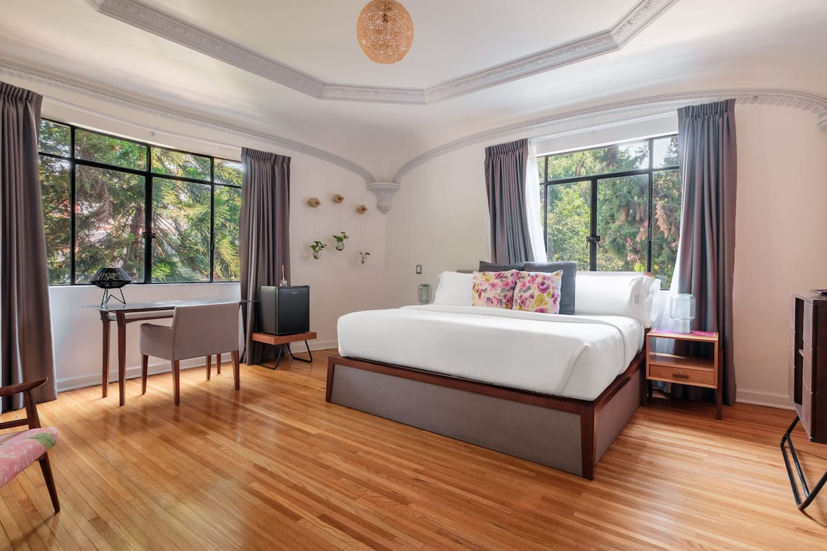 A spacious bedroom with a large bed, a desk, and a chair. There are two windows with curtains, a ceiling light, and a small table with a lamp.
