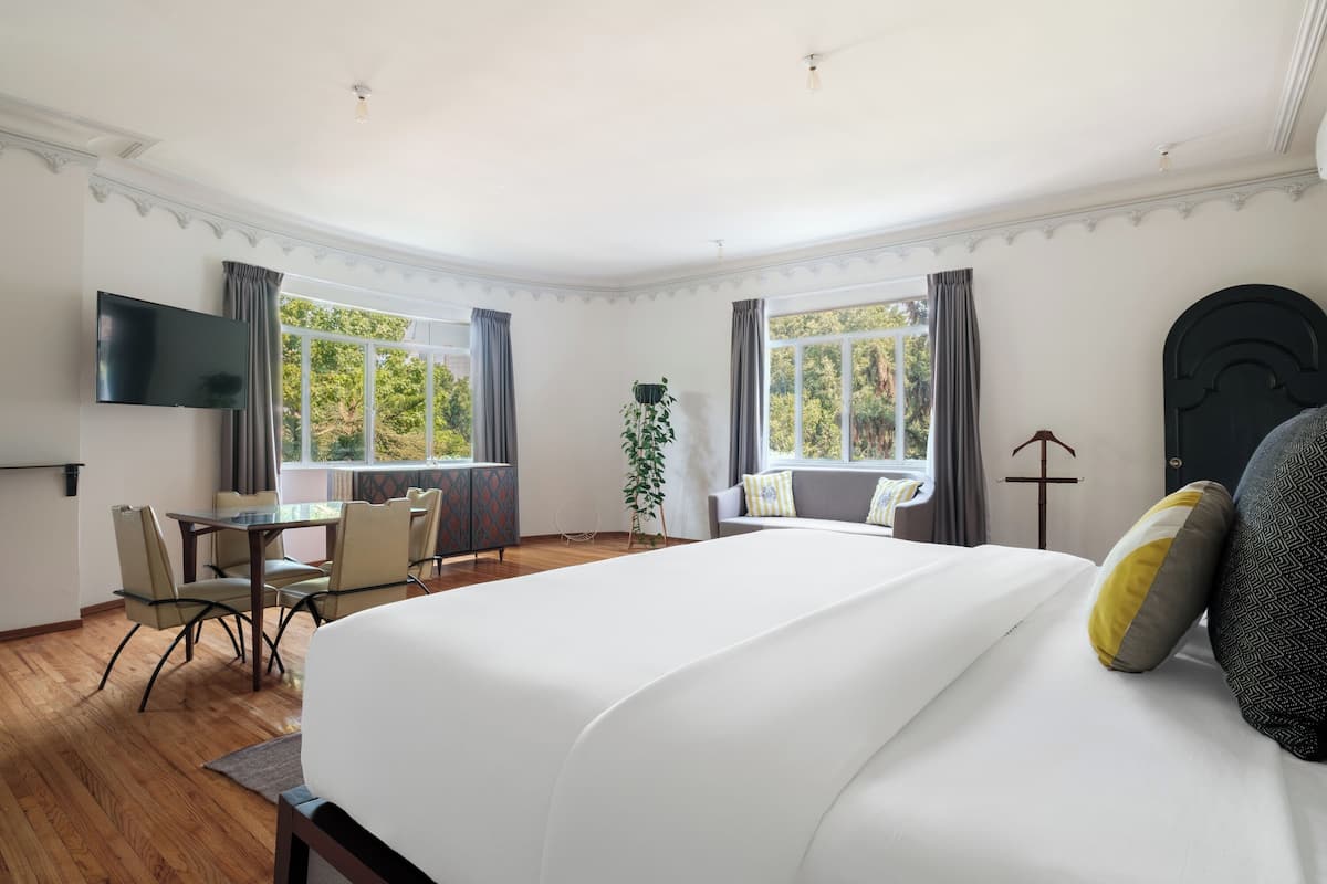 A hotel room with a large bed, a desk with chairs, a TV, and large windows with curtains.