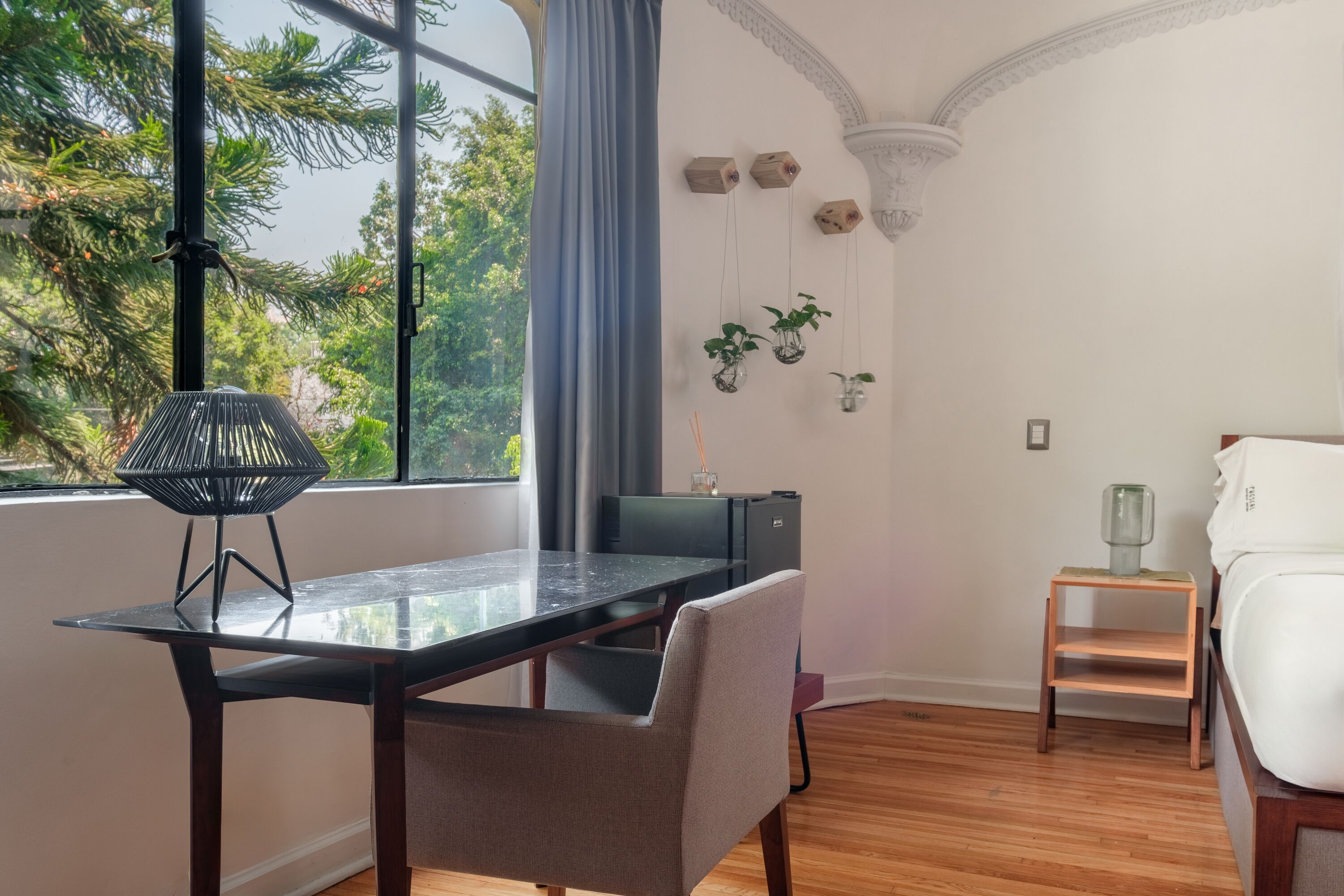 A modern room with a glass dining table, a grey chair, a small wooden side table, a white bed, and a view of trees through large windows.